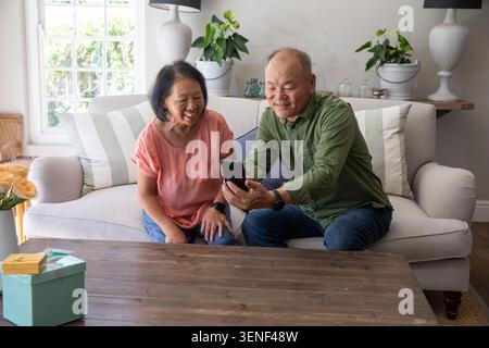 Donna cinese anziana e uomo cinese maturo seduto sul divano, condividendo smartphone e sorridendo Foto Stock