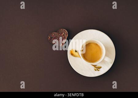 Tazza di caffè con cioccolato a forma di cuore su sfondo marrone scuro. Romantico concetto di pausa caffè. Stile piatto. Foto Stock