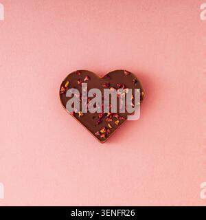 Composizione romantica di cioccolato a forma di cuore con petali di rosa secchi su sfondo rosa. Concetto d'amore minimalista. Stile piatto. Foto Stock