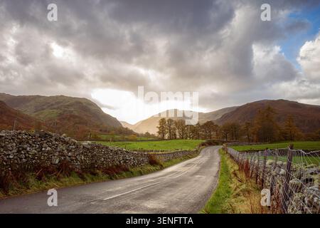 Una tranquilla strada rurale si snoda tra vecchie mura di pietra attraverso una valle verde nel Lake District, in Cumbria, con spettacolari nuvole di tempesta che si radunano sopra la testa. Foto Stock