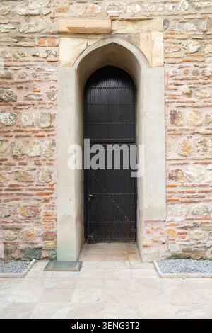 Skopje, Macedonia del Nord, 3 giugno 2026. Una porta di legno scuro e' posta all'interno di un arco di pietra in una parete di mattoni intemprati. L'area di fronte alla porta è pa Foto Stock