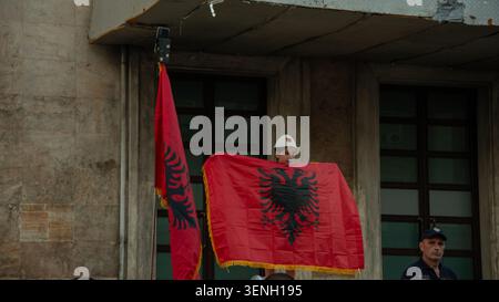 Il giovane dimostratore gilr tiene le bandiere nazionali albanesi da un balcone dell'edificio che si affaccia su una protesta anti-governativa a Tirana. Foto Stock