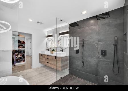 Bagno luminoso e contemporaneo con grande divisore di vetro che separa un'area doccia con piastrelle scure da un lavandino in legno chiaro con due specchi rotondi. OPE Foto Stock