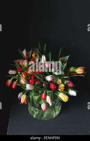 Tulipi colorati misti inclusi varietà di pappagallo in vaso di vetro, sfondo scuro, vivace composizione floreale primaverile Foto Stock
