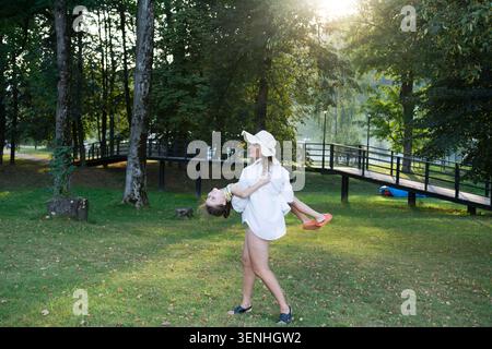 Una donna che indossa un cappello da sole e una camicia oversize fa oscillare giocosamente il suo bambino in un parco verdeggiante. La scena è immersa nel caldo sole del tardo pomeriggio Foto Stock