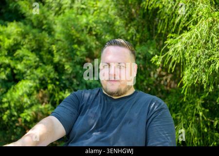 Giovane uomo seduto all'aperto in una calda giornata estiva con un'espressione premurosa e una postura informale. Uno stile di vita naturale che rappresenta relax, calore, l Foto Stock