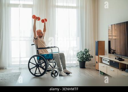 vista laterale dell'uomo in movimento in sedia a rotelle che fa esercizio con la pressa delle spalle in alto con manubri rossi in un luminoso soggiorno vicino alla tv che rappresenta il fi adattivo Foto Stock
