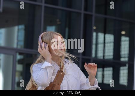 Donna in piedi di fronte a un edificio in vetro, che ascolta con le cuffie rosa e che tocca la tracolla della borsa Foto Stock