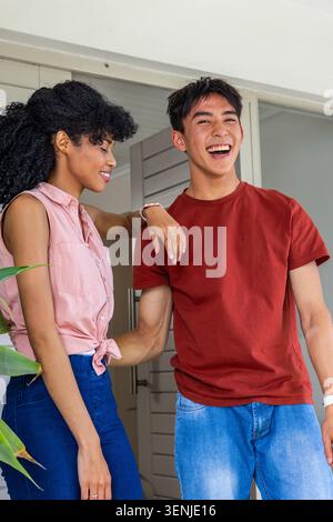 Donna afroamericana appoggiata all'uomo asiatico, ridendo sul portico con pianta in vaso e porta aperta Foto Stock