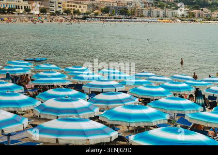 Sicilia, Italia, 20 giugno 2025 spiaggia affollata con file di ombrelloni blu e bianchi e persone che nuotano in mare in una soleggiata giornata estiva. ph di alta qualità Foto Stock