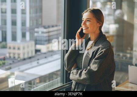 Felice giovane donna caucasica ventenne che parla al telefono cellulare vicino alla finestra dell'ufficio Foto Stock