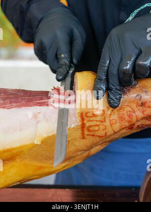 Primo piano di un prosciutto stagionato professionale tagliato a fette da una gamba di prosciutto con un coltello, tipico della salumeria europea, in Francia. Foto Stock