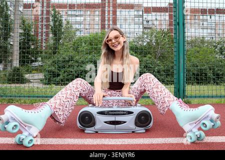 Sorridente giovane donna con pattini a rotelle e outfit retrò con stereo portatile su campi sportivi all'aperto, tiene un box tra le gambe, incarnando una spensieratezza Foto Stock
