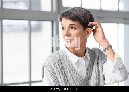 Donna matura e premurosa con capelli grigi corti e gilet in maglia a cavo che guarda lontano in un ambiente interno luminoso, indossa una camicia bianca con colletto sotto il tetto Foto Stock