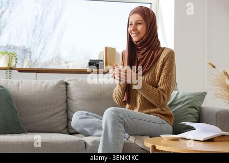 Donna musulmana sorridente in hijab che si rilassa sul divano con una tazza di caffè, si gode un'accogliente comodità domestica e il tempo libero, gode di un momento di comfort accogliente a casa Foto Stock