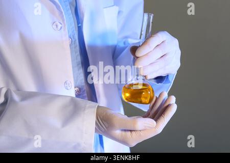 Scienziato in camice da laboratorio e guanti che reggono fiasca di vetro con liquido arancione per la ricerca chimica e la sperimentazione, l'attenzione è rivolta alle mani e alla f Foto Stock