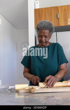 Donna anziana con baguette a fette sulla parte superiore verde su un tagliere di legno al banco della cucina con burro Foto Stock