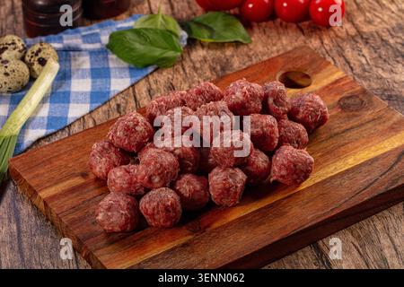 Un primo piano di polpette crude e crude disposte su un tagliere di legno scuro. Foglie di basilico verde fresco, pomodori rossi, uova di quaglia e un clo a scacchi Foto Stock