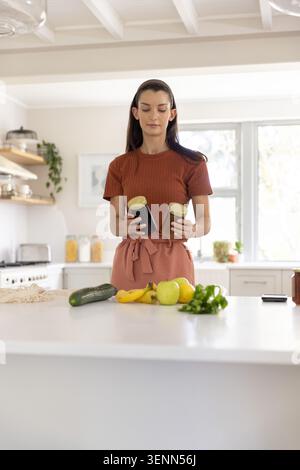 Donna di mezza età in piedi sull'isola bianca in grembiule rosa con due barattoli d'oro e prodotti Foto Stock