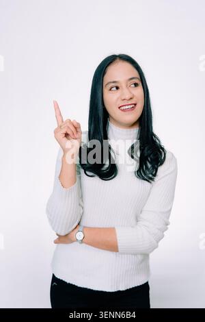 Giovane donna bruna indonesiana su sfondo bianco isolato che punta il dito verso lo spazio di copia, sorridendo e guardando di lato. Foto Stock