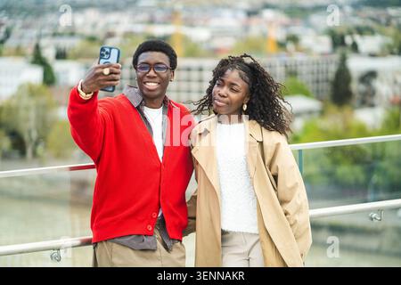 Felice afro-americano coppia di viaggiatori sposati che posano al punto panoramico della città facendo selfie al telefono. Foto Stock