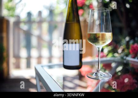 Una bottiglia di vino bianco con un'etichetta bianca si erge accanto a un bicchiere di vino riempito su un tavolo di vetro, circondato da un giardino con fiori e una recinzione di legno Foto Stock