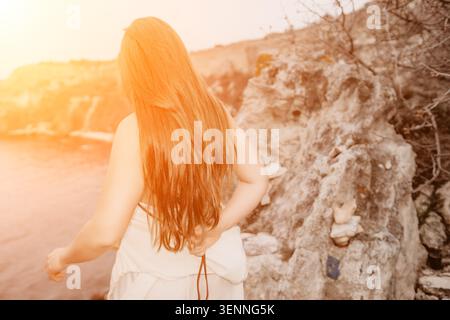 I capelli delle donne tramontano sulla costa rocciosa guardando il mare durante l'ora d'oro Foto Stock