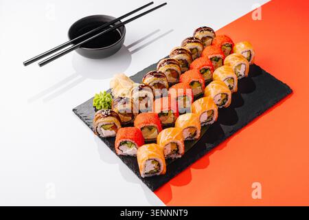 Gli involtini di sushi disposti ordinatamente su un piatto nero sono pronti da mangiare. Due tipi di rotoli sono dotati di guarnizioni colorate. Le bacchette e una piccola ciotola di salsa di soia si trovano nelle vicinanze. Foto Stock