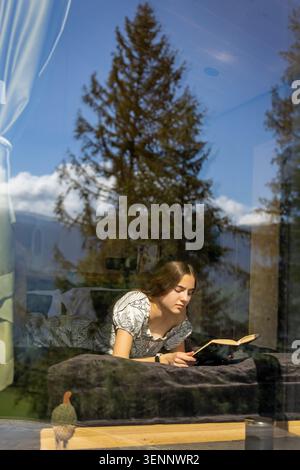 Donna che legge in cabina dietro la finestra della foresta riflettente. Una giovane donna giace su un letto scuro leggendo un libro aperto all'interno di una cabina di legno, vista attraverso Foto Stock