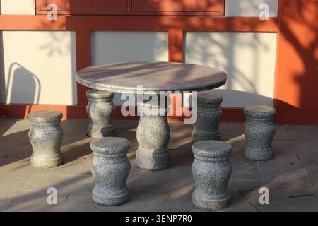 Tavolo e sedie in pietra si trovano sul patio esterno, proiettando ombre dalla luce del sole e dalle pareti arancioni. Foto Stock