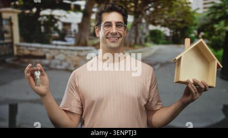 L'uomo tiene le chiavi con la mano sinistra e un modello di casa in legno con la mano destra su una strada, sorridendo e indossando occhiali; orgoglio di proprietà. Foto Stock