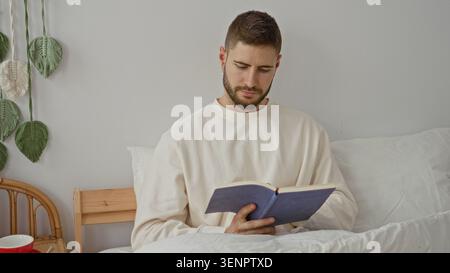 Uomo seduto a letto a leggere un libro cartaceo, mani che tengono le pagine aperte in una camera da letto con decorazioni a muro intrecciate e una tazza nelle vicinanze; riflesso tranquillo. Foto Stock