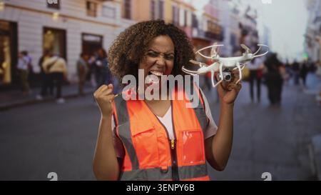 Donna con giubbotto di sicurezza arancione che tiene in mano il drone e fa una pompa a pugno su una strada trafficata della città con pedoni e vetrine sfocate; risultati celebrati Foto Stock