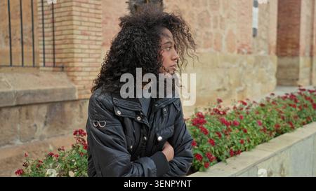 Teen Woman con i capelli ricci sta con le braccia incrociate accanto a un edificio in pietra e aiuole rosse; malinconica solitudine. Foto Stock