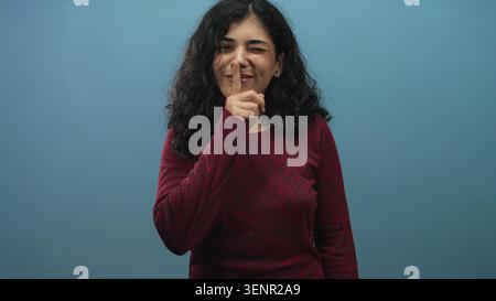 Donna araba che brilla dalle dita alle labbra in studio con sfondo blu con maglione rosso e un leggero sorriso; giocosa segretezza. Foto Stock