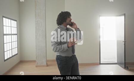 Uomo che tiene lo smartphone all'orecchio e punta il dito mentre si trova all'interno di un edificio vuoto; negoziazione mirata. Foto Stock