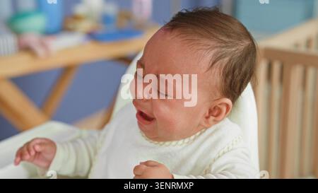 Bambino ispanico che piange con pugni stretti e striature di lacrime, rifiuta un biberon mentre è seduto in un seggiolone vicino alla culla e al fasciatoio; hun Foto Stock