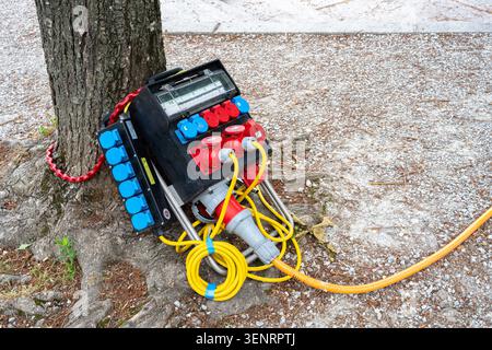 Losanna, Svizzera - 4 giugno 2026: Scatola di distribuzione dell'alimentazione esterna portatile con prese e cavi appoggiati su un albero su un terreno di ghiaia, utilizzata Foto Stock