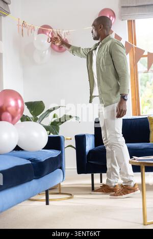 Il banner Happy Birthday in oro rosa è in fase di regolazione sulla parete del soggiorno, mentre i palloncini sono posizionati sul divano blu Foto Stock