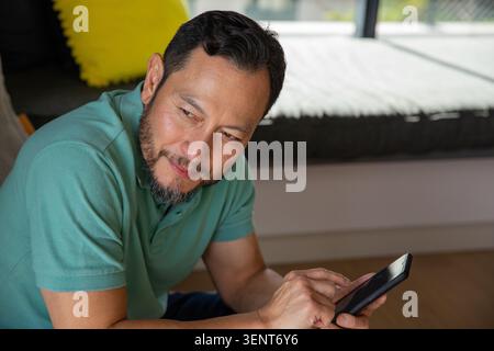 Uomo asiatico maturo seduto su panchina bassa vicino alla finestra, toccando telefono nero, polo verde acqua, cuscino giallo Foto Stock