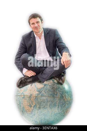 Il ritratto del dottor Gavin Francis è seduto su un grande globo. Indossa un abito e ha una postura rilassata. Lo sfondo è luminoso e semplice Foto Stock