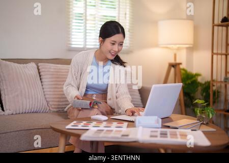 Giovane interior designer asiatico che lavora su progetti di design di mobili con campioni di materiale e computer nel soggiorno Foto Stock