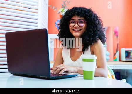 Giovane donna ispanica con capelli ricci e occhiali sorridenti alla telecamera, impegnata attivamente nel lavoro freelance o nello studio dalla sua comoda casa Foto Stock