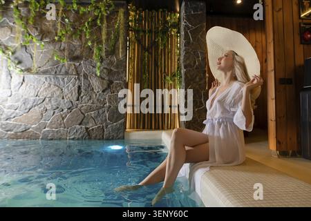 Una donna si rilassa in una lussuosa sauna, indossando un morbido accappatoio e un grande cappello da sole. L'atmosfera serena e' caratterizzata da calde tonalita' di legno e accenti di pietra, mentre Foto Stock