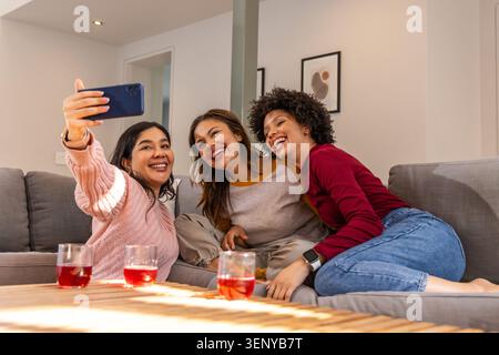 Diverse amiche che siedono sul divano grigio e scattano selfie con lo smartphone blu e tre bevande rosse Foto Stock