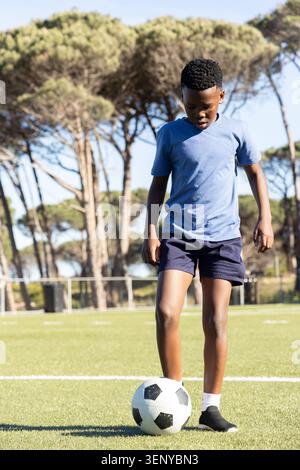 Bambino afroamericano maschio in piedi su un tappeto erboso con piede sulla palla indossando pantaloncini blu chiaro Foto Stock