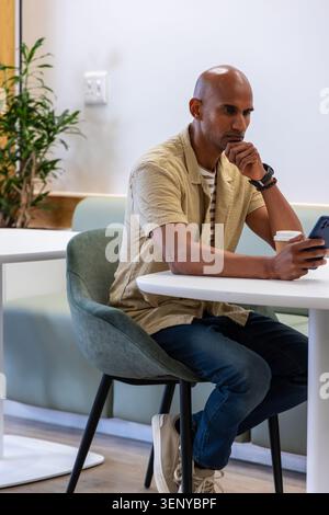 Uomo adulto seduto su una sedia nella caffetteria con indosso una camicia beige che tiene lo smartphone vicino alla tazza da caffè Foto Stock