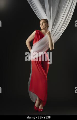 Una donna indossa con grazia un abito rosso che trasmette emozioni profonde. Interagisce con tessuti delicati drappeggiati in studio, sho Foto Stock