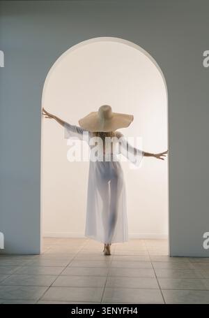 Questa immagine mostra una donna con una veste bianca e un cappello di paglia che cammina attraverso un grande arco bianco. I capelli delle donne sono biondi e lei ha le braccia allungate Foto Stock