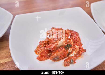 Contorno di gizzard fermentato asiatico ricoperto di pasta di peperoncino rosso ardente servito su un piatto di ceramica bianca su un tavolo di legno che mostra la sua cultura ora. Foto Stock